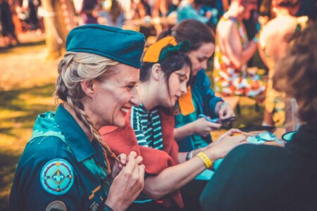 Women in patrol uniform and hat at the nipple tassel making activity at Camp Wildfire. A summer weekend break in a forest near London and Kent. An outdoor woodland retreat featuring adventure activities, live music, DJs, parties and camping. Half summer adventure activity camp, half music festival, for adults only.