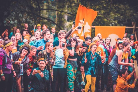 Crowd at the patrol games outside at Camp Wildfire. People representing Squirrel patrol at the Camp Wildfire patrol games. Girl in scarf with face paint on crawling through an obstacle at the Camp Wildfire patrol games. A summer weekend break in a forest near London and Kent. An outdoor woodland retreat featuring adventure activities, live music, DJs, parties and camping. Half summer adventure activity camp, half music festival, for adults only.