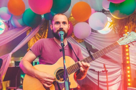 A member of the Keston Cobblers Club playing a guitar to the crowd in the discotheque tent at Camp Wildfire. A summer weekend break in a forest near London and Kent. An outdoor woodland retreat featuring adventure activities, live music, DJs, parties and camping. Half summer adventure activity camp, half music festival, for adults only.