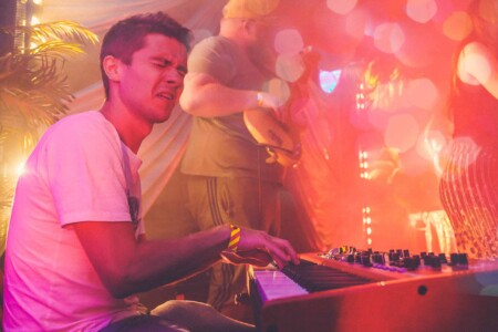 Man playing keyboard on stage at Camp Wildfire. A summer weekend break in a forest near London and Kent. An outdoor woodland retreat featuring adventure activities, live music, DJs, parties and camping. Half summer adventure activity camp, half music festival, for adults only.