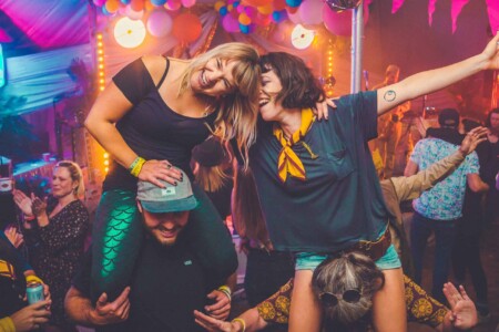 Girls on peoples shoulders in the discotheque tent at Camp Wildfire. A summer weekend break in a forest near London and Kent. An outdoor woodland retreat featuring adventure activities, live music, DJs, parties and camping. Half summer adventure activity camp, half music festival, for adults only.