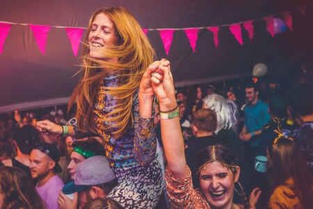 Girl with long ginger hair on someones shoulders in the discotheque tent at Camp Wildfire. A summer weekend break in a forest near London and Kent. An outdoor woodland retreat featuring adventure activities, live music, DJs, parties and camping. Half summer adventure activity camp, half music festival, for adults only.