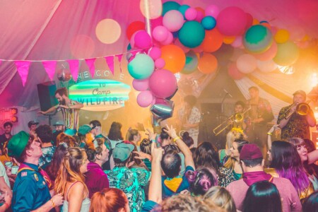 Colourfully dressed crowd in the decorated discotheque tent listening to a band at Camp Wildfire. A summer weekend break in a forest near London and Kent. An outdoor woodland retreat featuring adventure activities, live music, DJs, parties and camping. Half summer adventure activity camp, half music festival, for adults only.