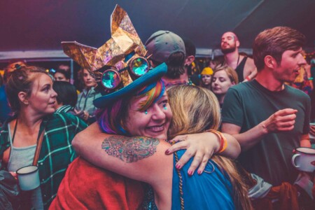 Lady in extravagant hat with colourful hair hugging a friend at Camp Wildfire. A summer weekend break in a forest near London and Kent. An outdoor woodland retreat featuring adventure activities, live music, DJs, parties and camping. Half summer adventure activity camp, half music festival, for adults only.