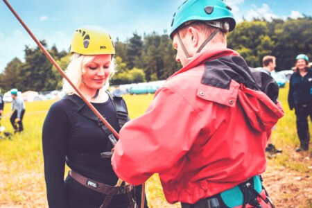 Blonde lady wearing a yellow helmet has her safety equipment checked. A summer weekend break in a forest near London and Kent. An outdoor woodland retreat featuring adventure activities, live music, DJs, parties and camping. Half summer adventure activity camp, half music festival, for adults only.