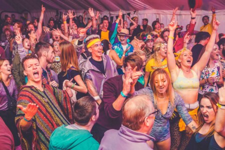 Large crowd throwing there hands up in the air in the discotheque tent at Camp Wildfire. A summer weekend break in a forest near London and Kent. An outdoor woodland retreat featuring adventure activities, live music, DJs, parties and camping. Half summer adventure activity camp, half music festival, for adults only.