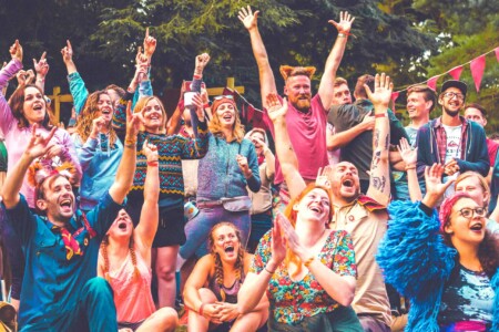 People dressed in colourful clothes cheering at the patrol games at Camp Wildfire. A summer weekend break in a forest near London and Kent. An outdoor woodland retreat featuring adventure activities, live music, DJs, parties and camping. Half summer adventure activity camp, half music festival, for adults only.