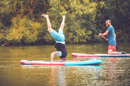 Man standing on his head at the stand up paddle boarding activity at Camp Wildfire. A summer weekend break in a forest near London and Kent. An outdoor woodland retreat featuring adventure activities, live music, DJs, parties and camping. Half summer adventure activity camp, half music festival, for adults only.