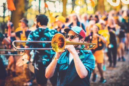 Adventurer in patrol uniform playing a trombone at Camp Wildfire. A summer weekend break in a forest near London and Kent. An outdoor woodland retreat featuring adventure activities, live music, DJs, parties and camping. Half summer adventure activity camp, half music festival, for adults only.