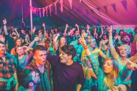 Crowd singing and partying in the discotheque tent at Camp Wildfire. A summer weekend break in a forest near London and Kent. An outdoor woodland retreat featuring adventure activities, live music, DJs, parties and camping. Half summer adventure activity camp, half music festival, for adults only.