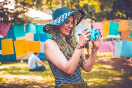 Girl in a floppy hat takes a photo using a blue camera. A summer weekend break in a forest near London and Kent. An outdoor woodland retreat featuring adventure activities, live music, DJs, parties and camping. Half summer adventure activity camp, half music festival, for adults only.