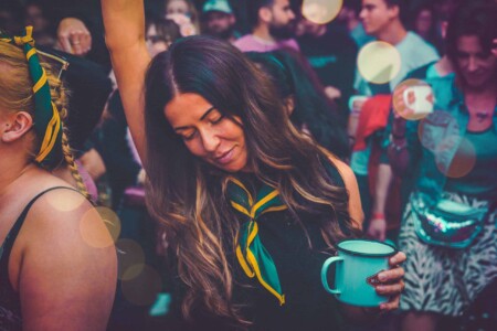 Girl with her patrol scarf on and branded enamel mug dancing at Camp Wildfire. A summer weekend break in a forest near London and Kent. An outdoor woodland retreat featuring adventure activities, live music, DJs, parties and camping. Half summer adventure activity camp, half music festival, for adults only.