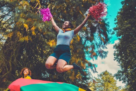 Girl in striped top jumping on trampoline with poms poms at Camp Wildfire. The UK's Best Alternative Festival. UK's Top Unique Event. A summer weekend break in a forest near London and Kent. An outdoor woodland retreat featuring adventure activities, live music, DJs, parties and camping. Half summer adventure activity camp, half music festival, for adults only.