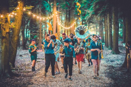 Adventurers in patrol uniforms with instruments marching in the Camp Wildfire parade. A summer weekend break in a forest near London and Kent. An outdoor woodland retreat featuring adventure activities, live music, DJs, parties and camping. Half summer adventure activity camp, half music festival, for adults only.