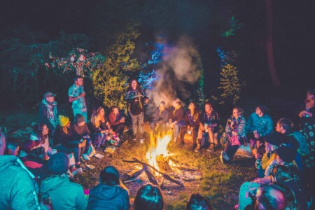 Woman storytelling round the campfire with friends at Camp Wildfire. The UK's Best Alternative Festival. UK's Top Unique Event. A summer weekend break in a forest near London and Kent. An outdoor woodland retreat featuring adventure activities, live music, DJs, parties and camping. Half summer adventure activity camp, half music festival, for adults only.