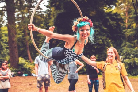 Blue haired lady hangs from hoop in forest. A summer weekend break in a forest near London and Kent. An outdoor woodland retreat featuring adventure activities, live music, DJs, parties and camping. Half summer adventure activity camp, half music festival, for adults only.